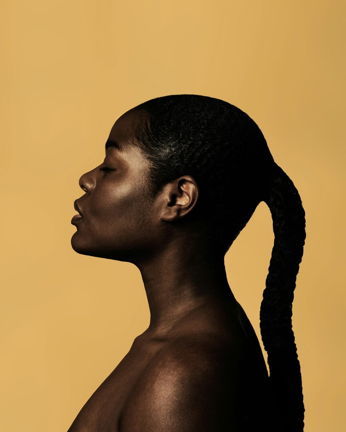 Side profile of a woman with a long braided ponytail and bare shoulders against a warm ochre backdrop